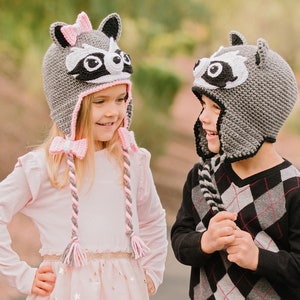 May include: Two children wearing gray raccoon hats with pink and black accents. The girl's hat has a pink bow. The boy's hat has a black and white striped tail. The children are smiling at each other.