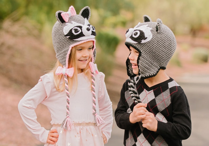May include: Two children wearing gray raccoon hats with pink and black accents. The girl's hat has a pink bow. The boy's hat has a black and white striped tail. The children are smiling at each other.