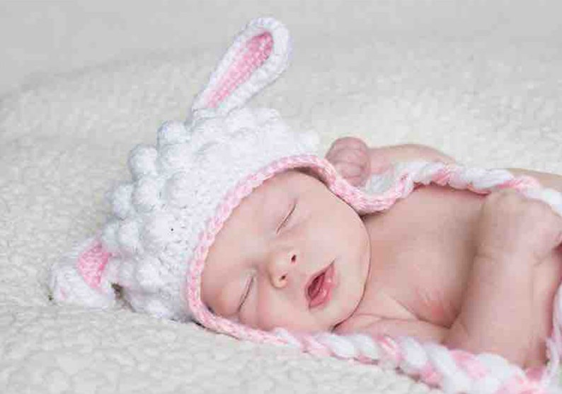 May include: A sleeping baby wearing a white and pink crochet bunny hat. The hat has long ears and a textured design. The baby is wrapped in a matching pink and white blanket.
