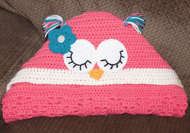 May include: A pink crocheted owl blanket with white and teal accents. The owl has white eyes with black eyelashes and a yellow beak. The blanket has a white crocheted border.