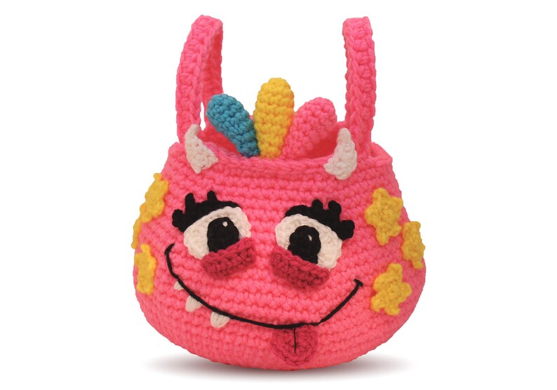 May include: Pink crocheted monster-shaped tote bag with a smiling face, white teeth, a pink tongue sticking out, and yellow stars. The bag has two white horns and a rainbow-colored crest on top.
