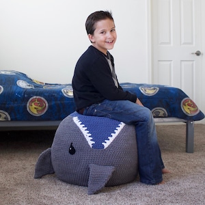 May include: A large, grey and blue crocheted whale-shaped ottoman. The ottoman has a blue seat with white teeth detailing. The whale has small fins and a black eye. A person is sitting on the ottoman.