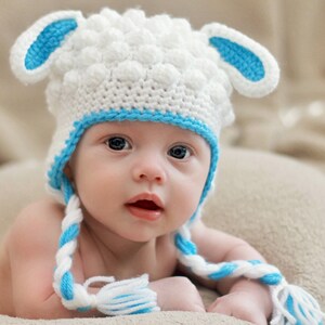 May include: A baby wearing a handmade white crochet hat with blue trim and ear details, resembling a sheep. The hat has braided ties with blue and white tassels. The baby is lying on a soft, neutral-colored blanket.