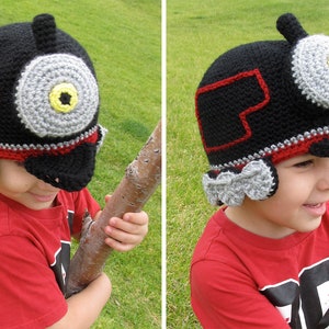 May include: A child wearing a black, red, and gray crocheted hat shaped like a train engine. The hat has a large, round, gray eye with a yellow center. The child is wearing a red shirt.