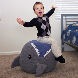 May include: A young boy sits on a large gray and blue crocheted shark-shaped beanbag chair. The chair has a white crocheted mouth with teeth and a black eye.
