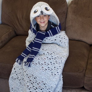May include: A child wearing a gray crocheted blanket with a white owl hood and a blue and white striped scarf. The child is sitting on a brown couch.