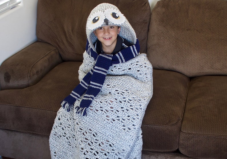 May include: A child wearing a gray crocheted blanket with a white owl hood and a blue and white striped scarf. The child is sitting on a brown couch.