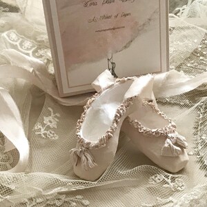 May include: A pair of off-white ballet slippers with ruffled edges and small bows, resting on a bed of lace and ribbons. A box with text is in the background. The slippers are a soft, muted color.