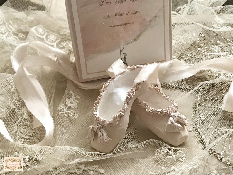 May include: A pair of off-white ballet slippers with ruffled edges and small bows, resting on a bed of lace and ribbons. A box with text is in the background. The slippers are a soft, muted color.
