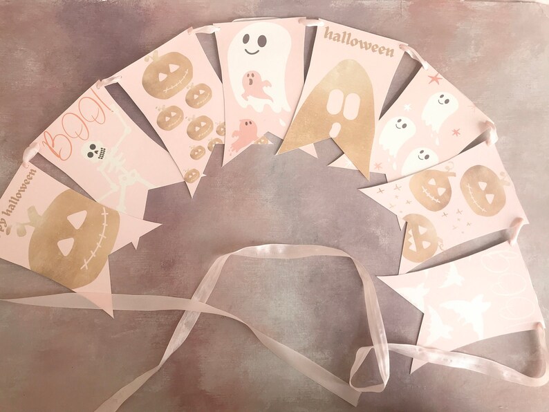 May include: A decorative bunting banner featuring pale pink pennants strung on a ribbon. The flags display gold and white Halloween-themed graphics including pumpkins, ghosts, a skeleton, and the text BOO! and halloween in various fonts.