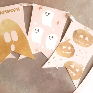 May include: A decorative banner featuring pale pink pennants strung together with ribbon. The pennants display gold foil graphics of a large ghost, three white ghosts with stars, and three gold foil jack-o'-lanterns. The word halloween is printed in gold.