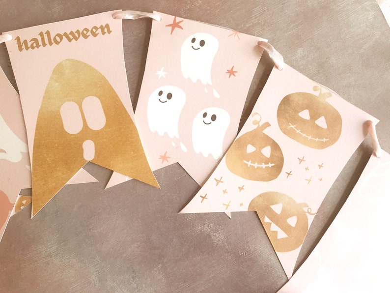 May include: A decorative banner featuring pale pink pennants strung together with ribbon. The pennants display gold foil graphics of a large ghost, three white ghosts with stars, and three gold foil jack-o'-lanterns. The word halloween is printed in gold.