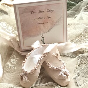 May include: A pair of handmade ballet slippers in a soft, off-white hue, adorned with delicate ruffles and bows. The slippers are displayed with a box that reads "Tora Pearl Design" and "A Muse of Paper."