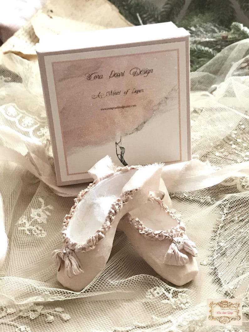 May include: A pair of handmade ballet slippers in a soft, off-white hue, adorned with delicate ruffles and bows. The slippers are displayed with a box that reads "Tora Pearl Design" and "A Muse of Paper."
