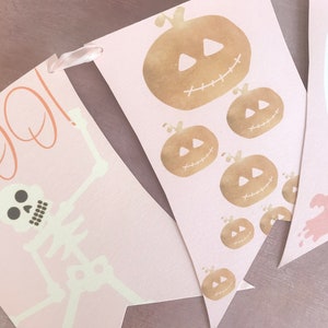 May include: A decorative banner featuring three pennants in a soft pink hue. The first pennant displays a skeleton and the text BOO in orange. The second shows multiple gold-toned pumpkins. The third features white and pink ghost illustrations.