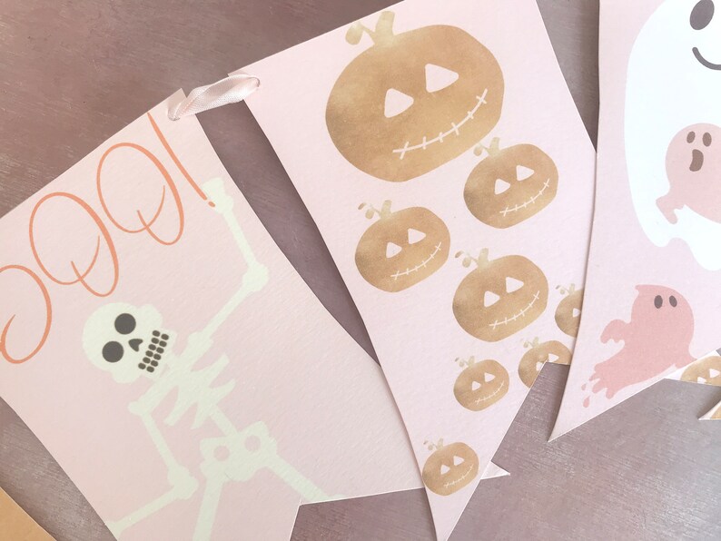 May include: A decorative banner featuring three pennants in a soft pink hue. The first pennant displays a skeleton and the text BOO in orange. The second shows multiple gold-toned pumpkins. The third features white and pink ghost illustrations.