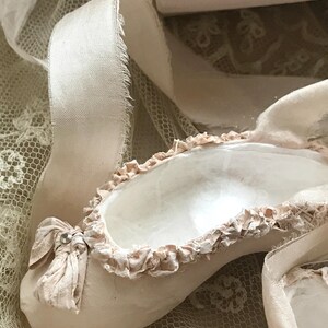 May include: A close-up of a ballet slipper in a soft, muted pink hue. The slipper features a delicate bow with a small jewel and is trimmed with ruffled fabric. A cream-colored ribbon and lace fabric are also visible.