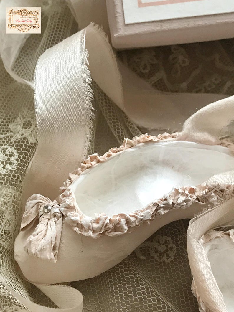 May include: A close-up of a ballet slipper in a soft, muted pink hue. The slipper features a delicate bow with a small jewel and is trimmed with ruffled fabric. A cream-colored ribbon and lace fabric are also visible.