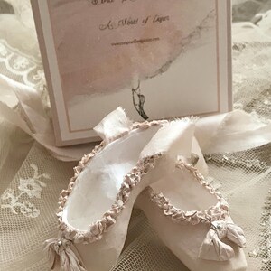 May include: A pair of delicate, off-white ballet slippers with ruffled edges and small bows. The slippers are displayed on a lace surface, with a box in the background that reads "Tora Pearl Design" and "A Moment of Luster."