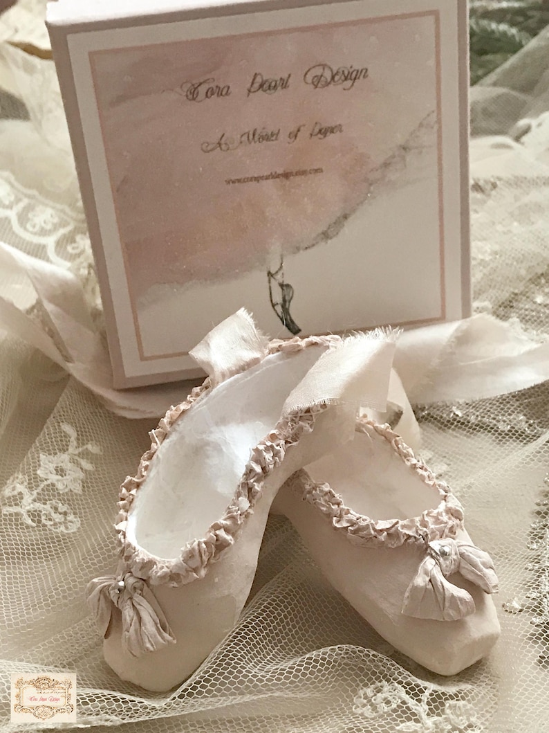 May include: A pair of delicate, off-white ballet slippers with ruffled edges and small bows. The slippers are displayed on a lace surface, with a box in the background that reads "Tora Pearl Design" and "A Moment of Luster."