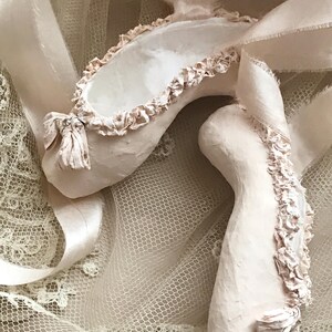 May include: A pair of pale pink ballet slippers with delicate ruffled edges and satin ribbons. The slippers have a bow detail with a small embellishment. The background is a soft, textured fabric.