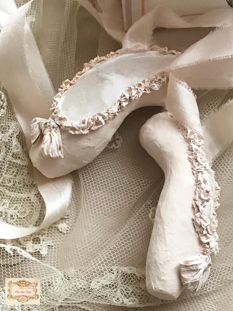May include: A pair of pale pink ballet slippers with delicate ruffled edges and satin ribbons. The slippers have a bow detail with a small embellishment. The background is a soft, textured fabric.