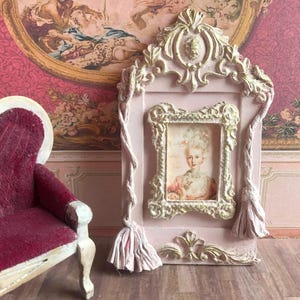 May include: A miniature pink framed picture with a portrait of a woman in a decorative gold frame. The frame is embellished with gold accents and tassels. A small burgundy sofa is to the left of the frame.