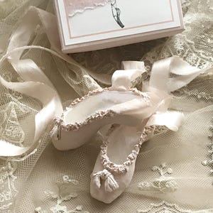 May include: A pair of off-white ballet slippers with delicate ribbon ties, adorned with ruffled trim and small bows. The slippers rest on a cream-colored lace fabric, with a small box in the background. The box has a pink and white design and the text "www.uniquepaperdesigns.etsy.com".