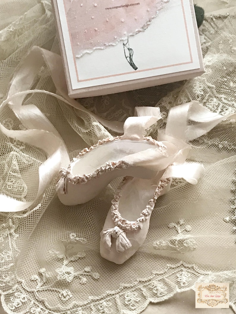May include: A pair of off-white ballet slippers with delicate ribbon ties, adorned with ruffled trim and small bows. The slippers rest on a cream-colored lace fabric, with a small box in the background. The box has a pink and white design and the text "www.uniquepaperdesigns.etsy.com".
