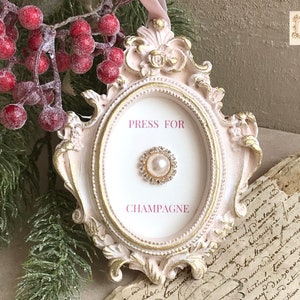 May include: A decorative, oval-shaped frame in white and gold, with the text "PRESS FOR CHAMPAGNE." The frame features a pearl and rhinestone centrepiece. The image also shows red berries and foliage.