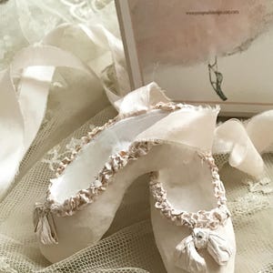 May include: A pair of off-white ballet slippers with ruffled trim and ribbon ties. The slippers are displayed on a lace surface, with a decorative card in the background that reads "Tora Pearl Design".