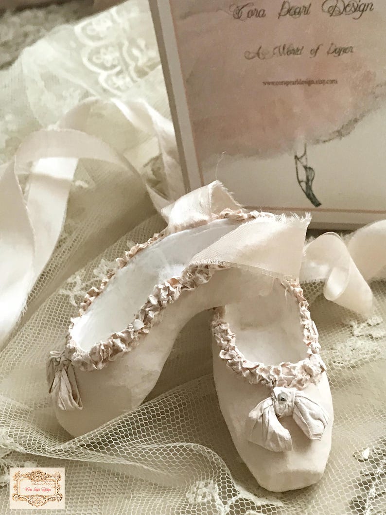 May include: A pair of off-white ballet slippers with ruffled trim and ribbon ties. The slippers are displayed on a lace surface, with a decorative card in the background that reads "Tora Pearl Design".