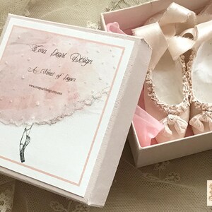 May include: A pair of pale pink ballet slippers with delicate bows and ruffled edges, presented in an open, light pink box. The box features a watercolor-style design and the text "Tora Pearl Design".
