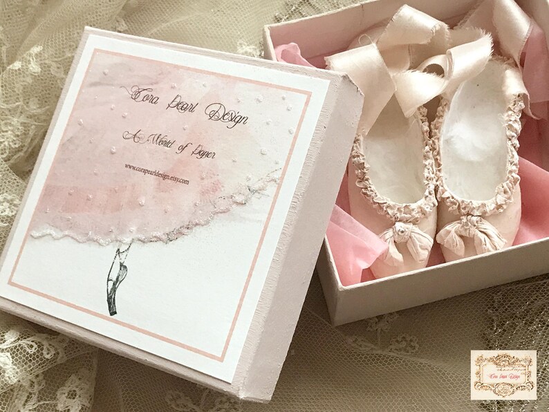 May include: A pair of pale pink ballet slippers with delicate bows and ruffled edges, presented in an open, light pink box. The box features a watercolor-style design and the text "Tora Pearl Design".