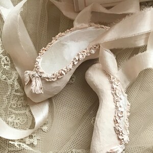 May include: A pair of off-white ballet slippers with ruffled edges and ribbon ties. The slippers are placed on a cream-colored lace surface, with the ribbons gracefully draped around them. The overall aesthetic is soft and delicate.