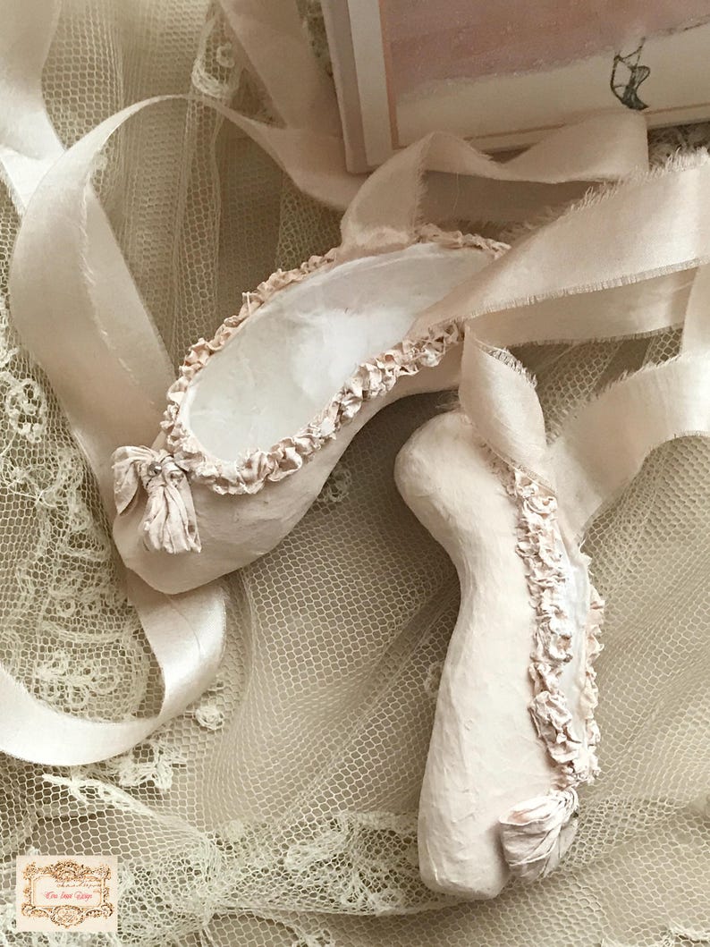 May include: A pair of off-white ballet slippers with ruffled edges and ribbon ties. The slippers are placed on a cream-colored lace surface, with the ribbons gracefully draped around them. The overall aesthetic is soft and delicate.