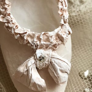 May include: Close-up of a ballet slipper in a soft pink hue. The slipper features a delicate bow with a sparkling embellishment and a textured, ruffled trim around the opening. The overall design is elegant and feminine.