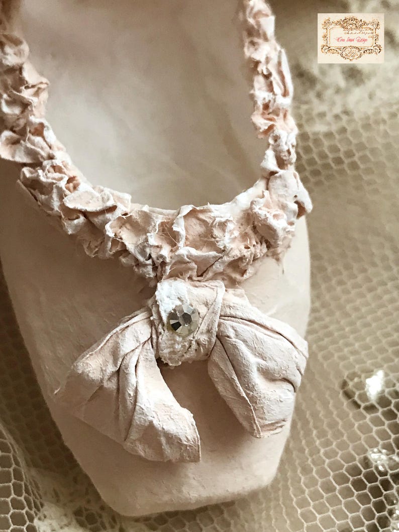 May include: Close-up of a ballet slipper in a soft pink hue. The slipper features a delicate bow with a sparkling embellishment and a textured, ruffled trim around the opening. The overall design is elegant and feminine.