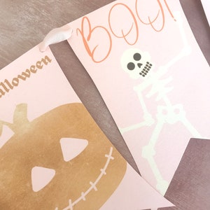May include: A decorative banner featuring pale pink pennants strung together with white ribbon. One pennant displays a gold pumpkin with the text "ppy halloween" and another shows a white skeleton with the orange text "BOO!" above it.