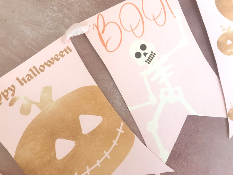 May include: A decorative banner featuring pale pink pennants strung together with white ribbon. One pennant displays a gold pumpkin with the text "ppy halloween" and another shows a white skeleton with the orange text "BOO!" above it.