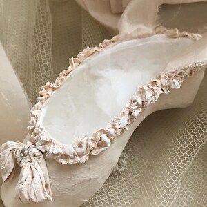 May include: A single, handmade ballet slipper in a soft, muted pink hue. The slipper features a ruffled trim around the opening and a delicate bow detail at the toe. The background includes a sheer, net-like fabric and ribbons, enhancing the delicate aesthetic.