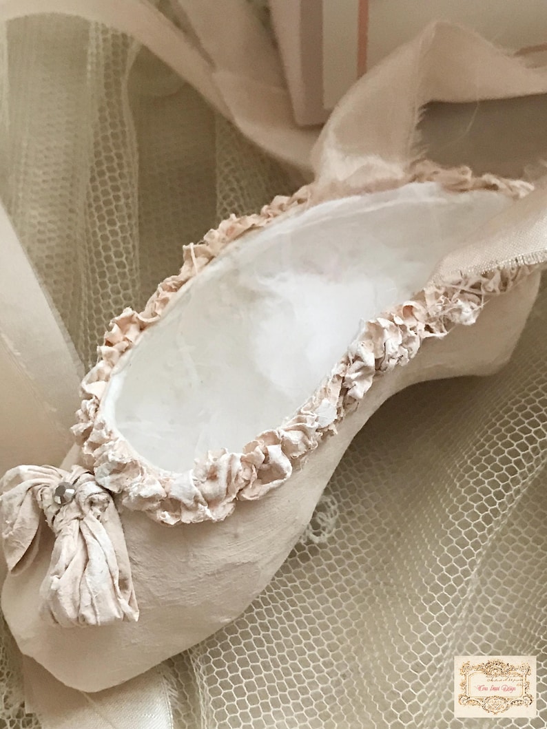 May include: A single, handmade ballet slipper in a soft, muted pink hue. The slipper features a ruffled trim around the opening and a delicate bow detail at the toe. The background includes a sheer, net-like fabric and ribbons, enhancing the delicate aesthetic.