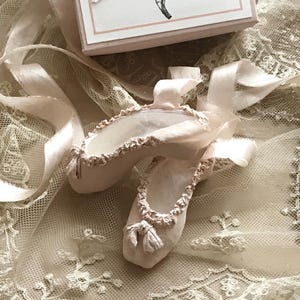 May include: A pair of pale pink ballet slippers with satin ribbons, resting on a delicate lace surface. The slippers have ruffled edges and small bows at the toes. A small box with a ballerina illustration is in the background.
