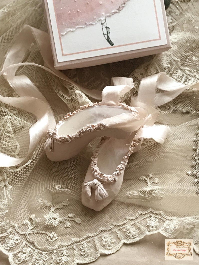 May include: A pair of pale pink ballet slippers with satin ribbons, resting on a delicate lace surface. The slippers have ruffled edges and small bows at the toes. A small box with a ballerina illustration is in the background.