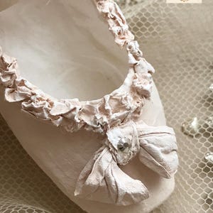 May include: A close-up of a pale pink ballet slipper with a ruffled trim and a bow. The bow is adorned with a small, sparkly embellishment. The slipper is set against a soft, textured background.