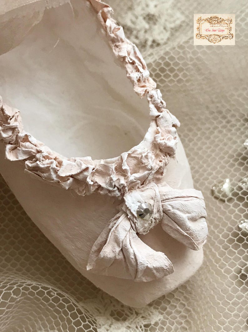 May include: A close-up of a pale pink ballet slipper with a ruffled trim and a bow. The bow is adorned with a small, sparkly embellishment. The slipper is set against a soft, textured background.