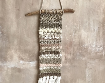 Puede incluir: Un tapiz de pared tejido con una estética natural. La pieza presenta una variedad de texturas y colores, incluyendo beige, crema y marrón. Se suspende de una varilla de madera con una cuerda crema y tiene flecos en la parte inferior. Mide unos 40 cm.