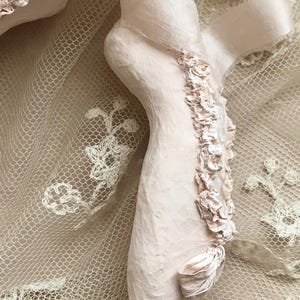 May include: A pale pink plaster foot sculpture with delicate fabric accents. The foot is adorned with a ribbon and small, ruffled fabric flowers. The sculpture rests on a backdrop of cream-colored lace and ribbon.