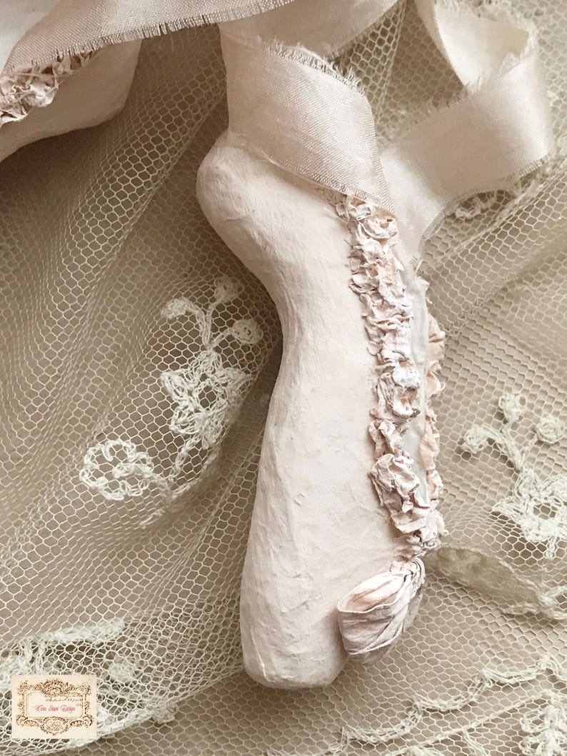May include: A pale pink plaster foot sculpture with delicate fabric accents. The foot is adorned with a ribbon and small, ruffled fabric flowers. The sculpture rests on a backdrop of cream-colored lace and ribbon.