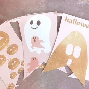 May include: A decorative banner featuring light pink pennants strung on a ribbon. The pennants display gold foil jack-o'-lanterns, white ghosts with smiling faces, smaller pink ghosts, and a gold foil ghost shape with the text halloween.